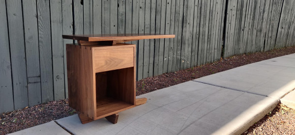 A walnut nightstand with a slab top that cantilevers to the right of a one-drawer carcass.